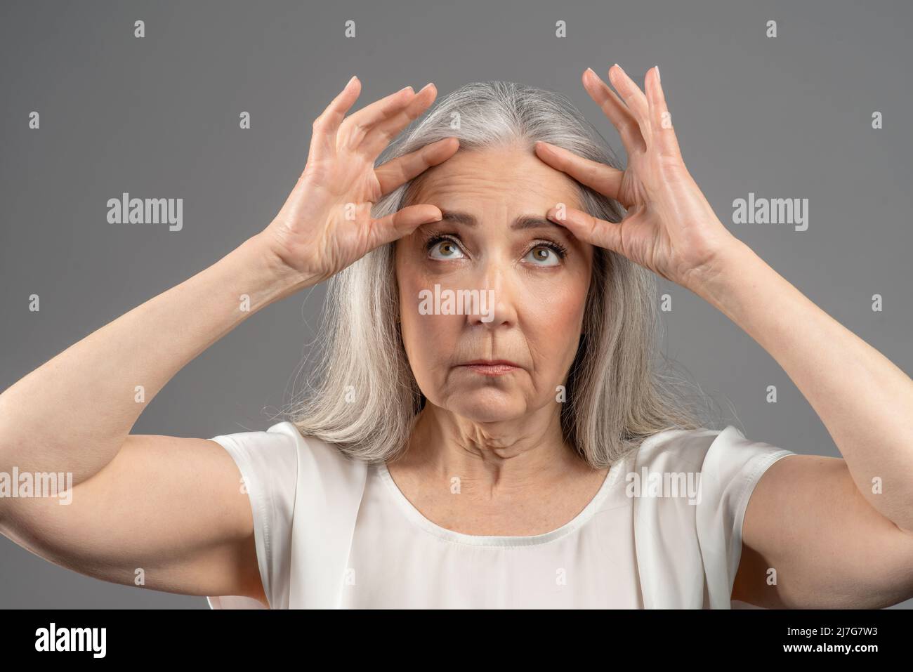 Porträt einer älteren Frau, die mit Falten auf ihrer Stirn unzufrieden ist und das Gesicht auf grauem Studiohintergrund berührt Stockfoto