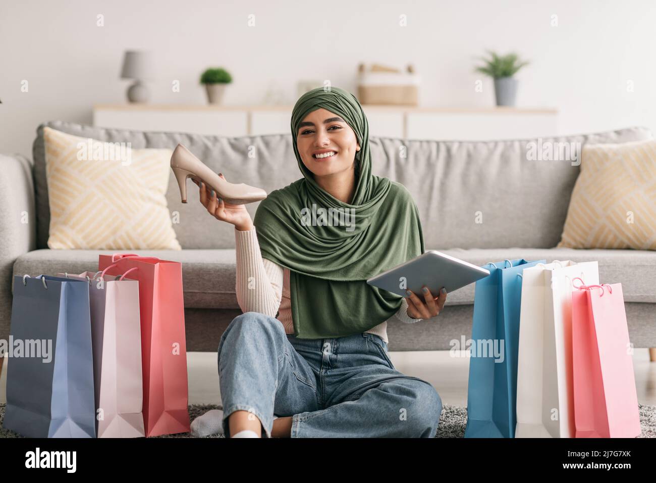 Junge arabische Frau in Hijab hält digitales Tablet und neuen Schuh, sitzt auf dem Boden zwischen Geschenktüten, schreiben Feedback Bewertung Stockfoto