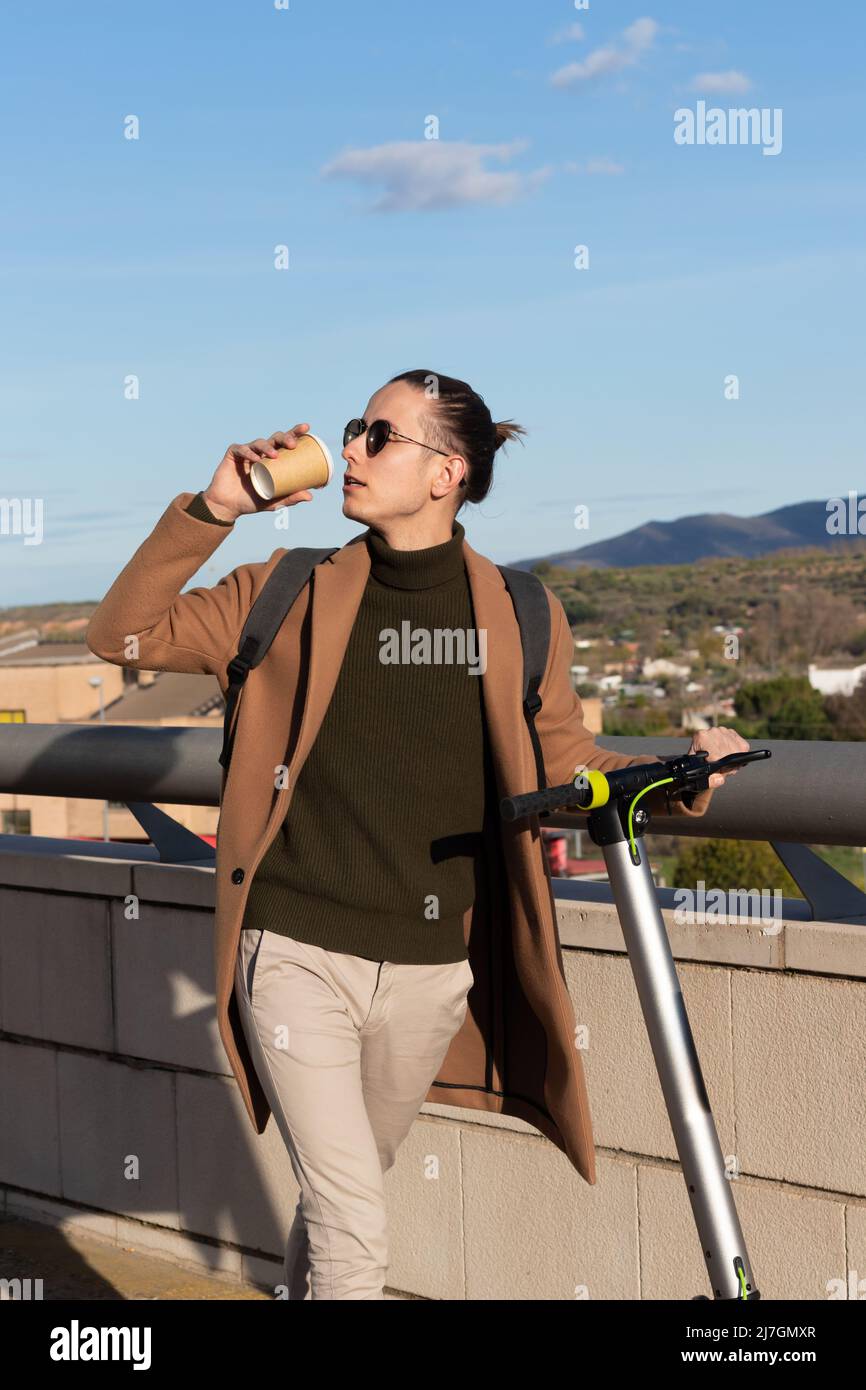 Vertikale Nahaufnahme eines jungen, hübschen Mannes mit Sungalasses, Elektroroller und einer Tasse Kaffee unter blauem Himmel und Berglandschaft Stockfoto