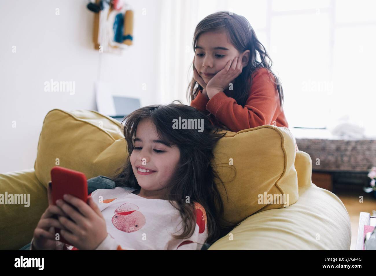 Mädchen mit Kopf in den Händen, die Schwester mit dem Telefon ansieht, während sie auf dem Sessel im Wohnzimmer sitzt Stockfoto