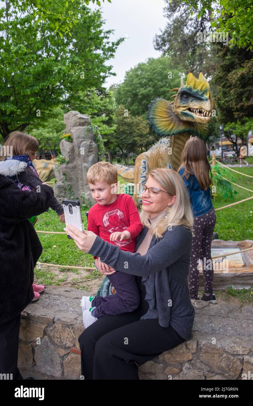 Mutter hält ihren Sohn in den Armen und nimmt ein Selfie mit ihrem Handy beim Dinosaurierfest. Stockfoto