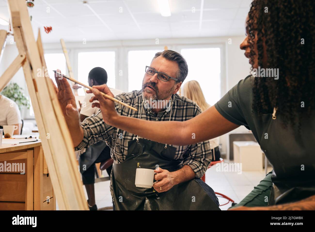 Männlicher Tutor, der weiblichen Studenten bei der Malerei auf der Leinwand des Künstlers in der Kunstklasse assistiert Stockfoto