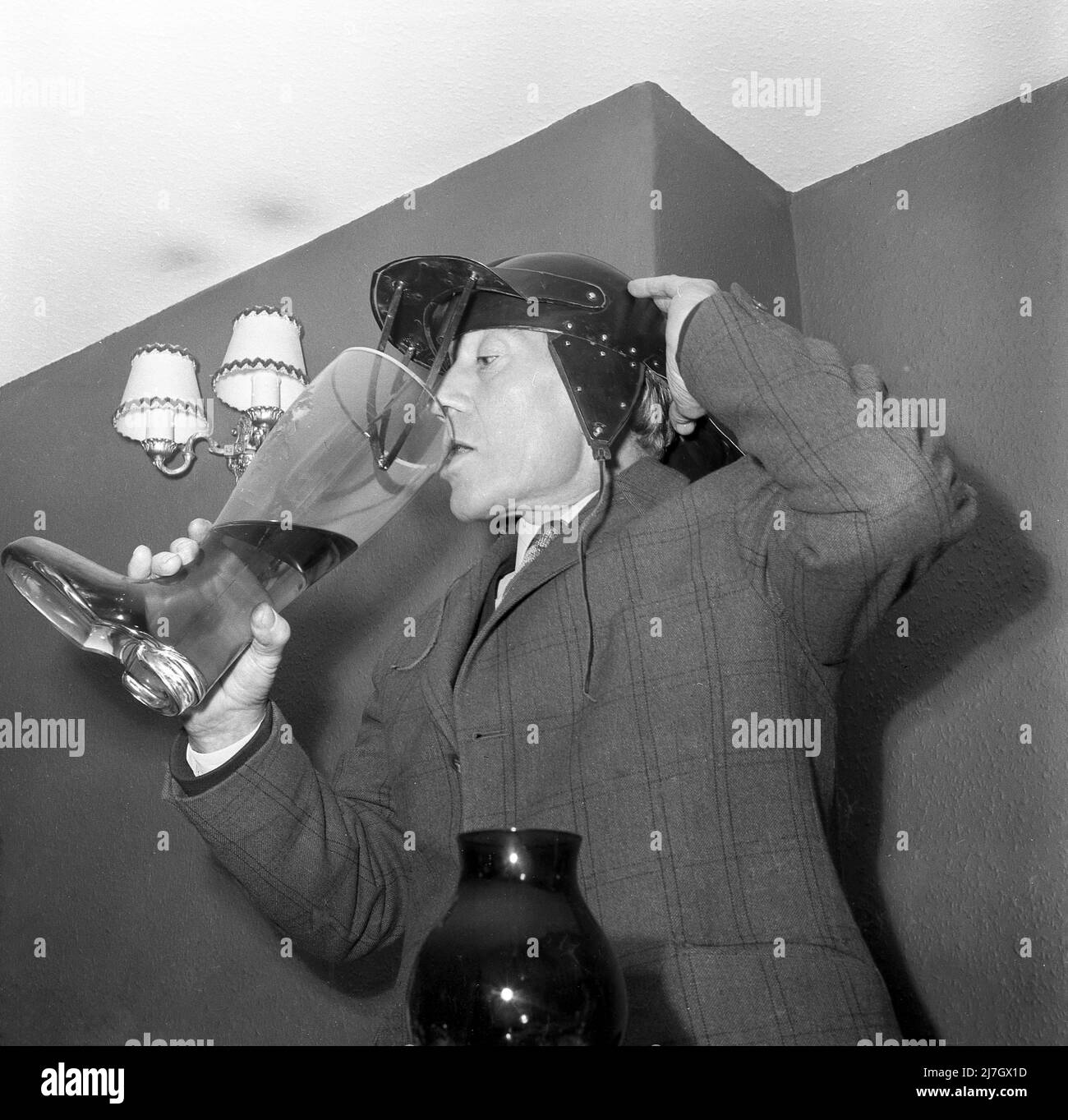 1960s, historisch, ein Mann, der in einem Raum steht, eine Jacke mit Karokmuster und eine ungewöhnliche Mütze oder einen Hut mit Ohrenklappen trägt. Der Hut hat auch eine Front-Visier mit anliegenden Lederriemen. Er trinkt von einem großen Glas Bier Stiefel, vielleicht ein traditioneller Toast oder Ritual bei einem jährlichen Sportessen. In Deutschland ist das Biertrinken aus einem Glasstiefel eine Tradition aus dem 19.. Jahrhundert, wo es als militärisches Ritual für junge Soldaten begann. Stockfoto