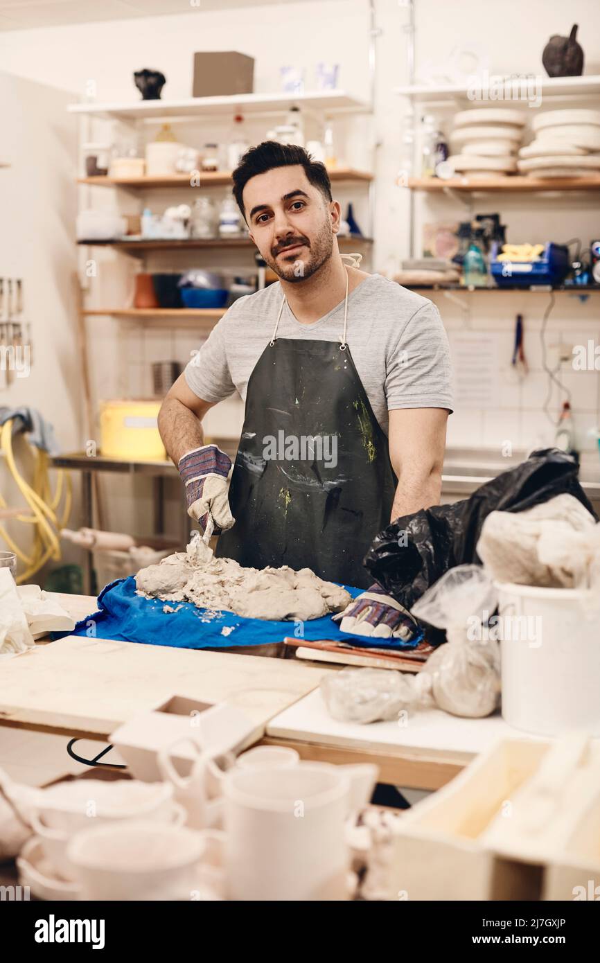 Junger Mann mit Schürze, der mit Ton am Tisch in der Kunstklasse stand Stockfoto