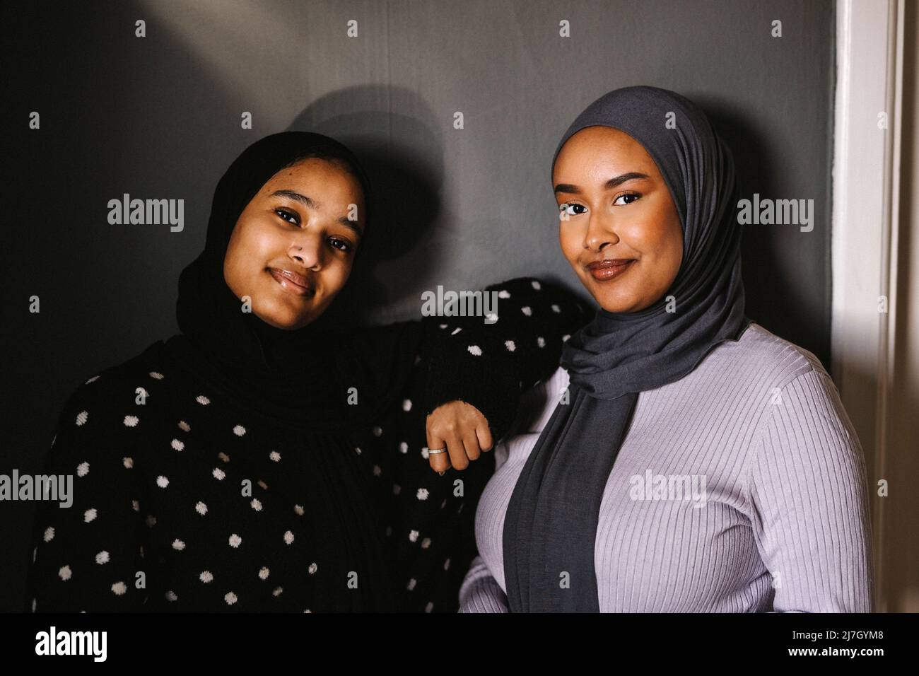 Lächelnde junge Frauen in Hijabs, die zu Hause vor der Wand stehen Stockfoto