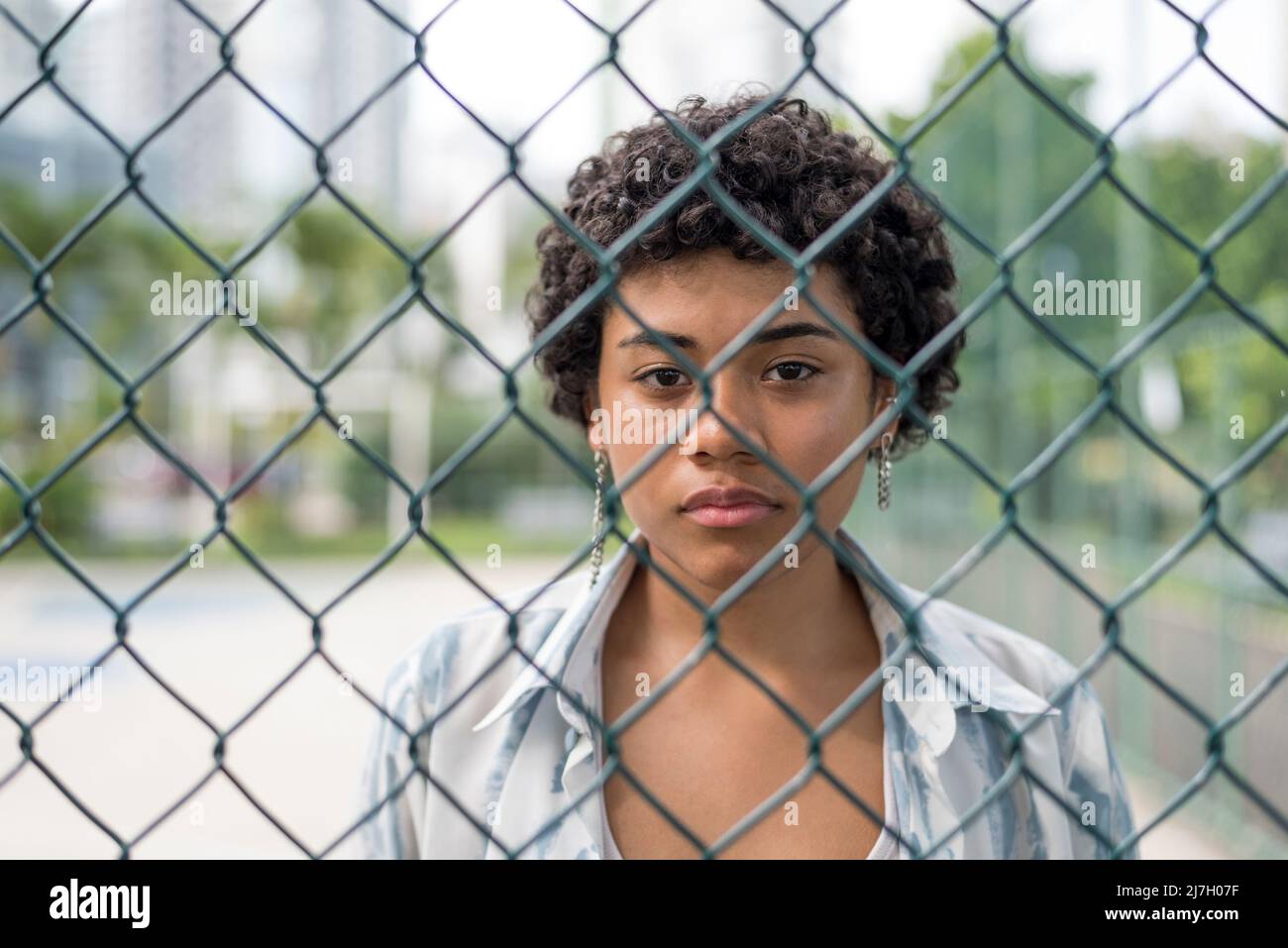 Nahaufnahme einer jungen lateinerin, die mit ernstem Gesicht auf die Kamera schaut, während sie hinter einem gewebten Drahtzaun im Park steht. Stockfoto