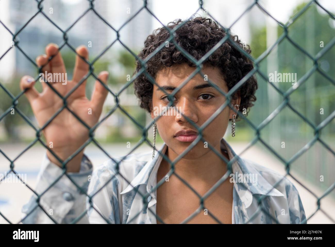 Nahaufnahme einer jungen lateinerin, die mit ernstem Gesicht auf die Kamera schaut, während sie hinter einem gewebten Drahtzaun im Park steht. Stockfoto