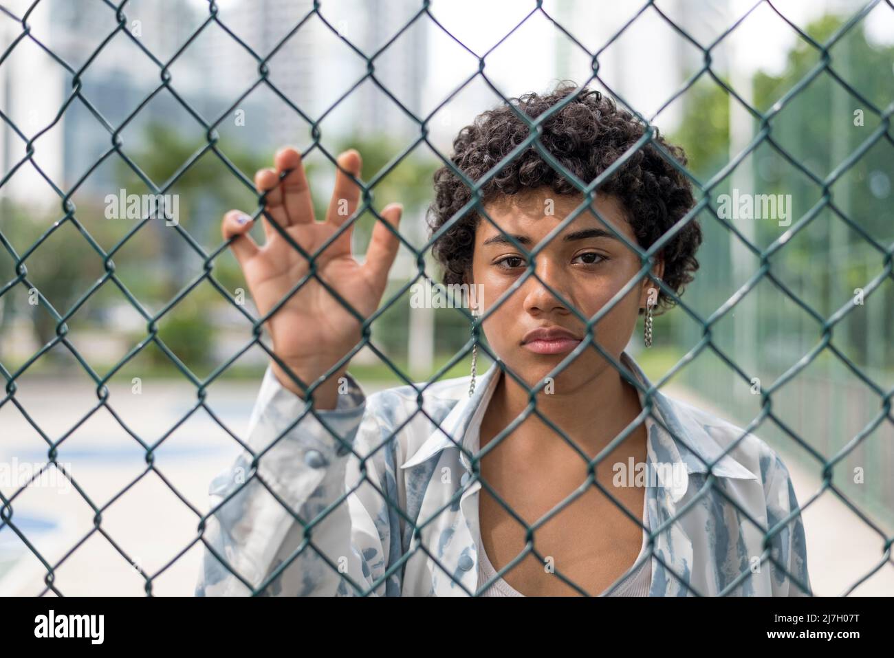 Nahaufnahme einer jungen lateinerin, die mit ernstem Gesicht auf die Kamera schaut, während sie hinter einem gewebten Drahtzaun im Park steht. Stockfoto