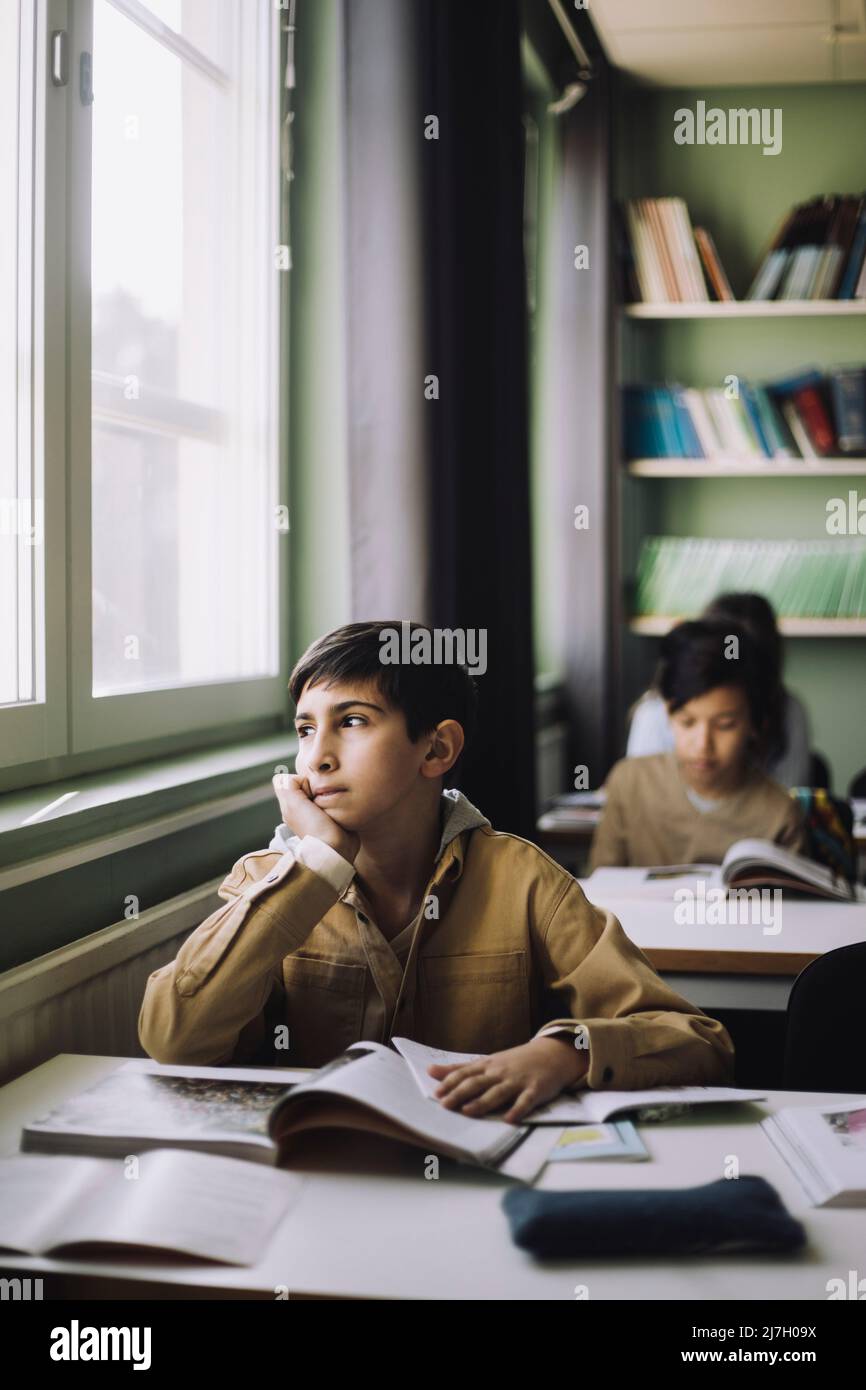 Nachdenklicher Junge mit der Hand auf dem Kinn, der im Klassenzimmer vor dem Fenster schaute Stockfoto