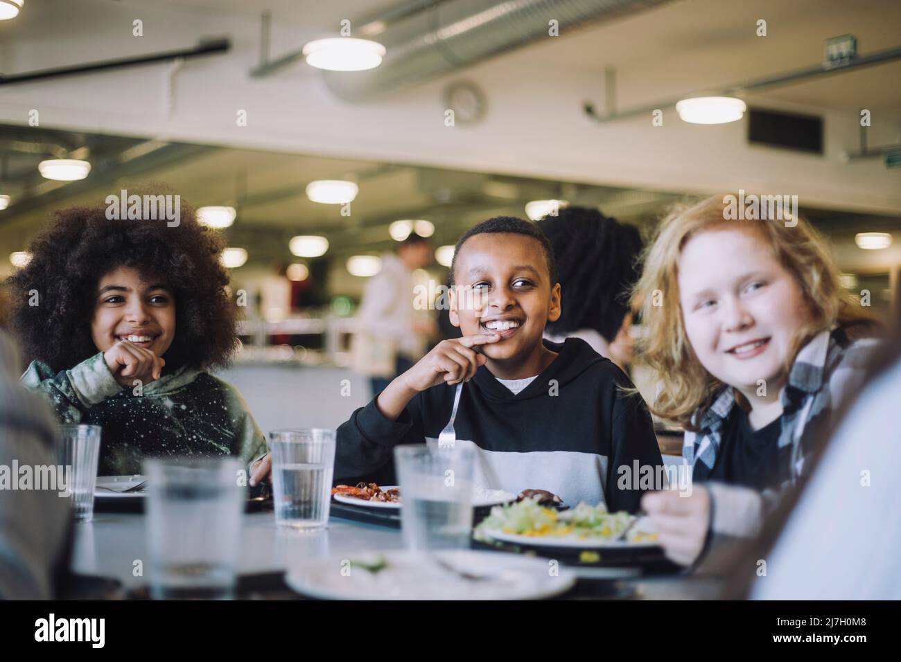 Lächelnde Schüler blicken weg, während sie während der Mittagspause in der Cafeteria am Tisch sitzen Stockfoto