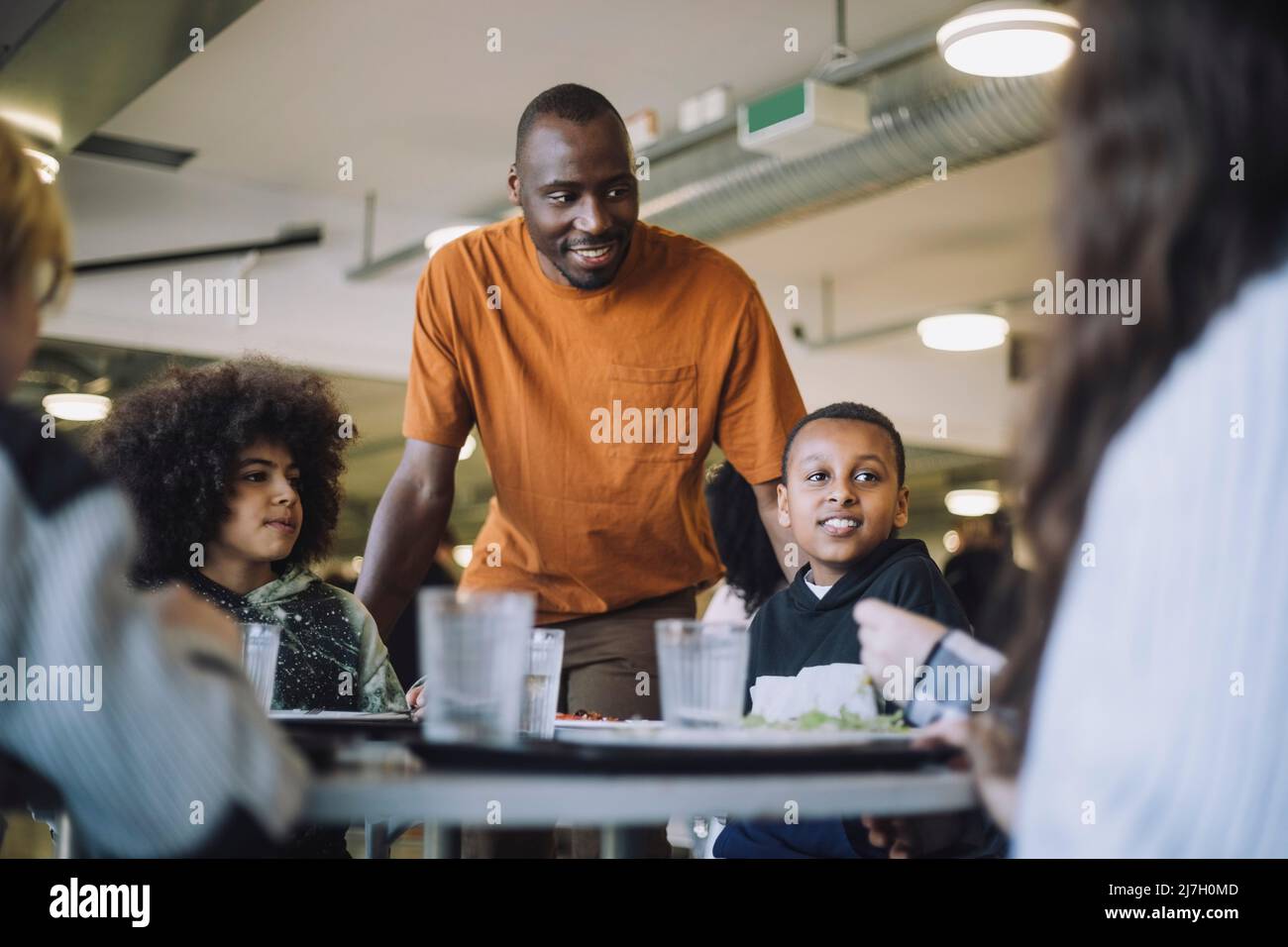 Lächelnder Lehrer, der während der Mittagspause in der Cafeteria bei den Schülern stand Stockfoto