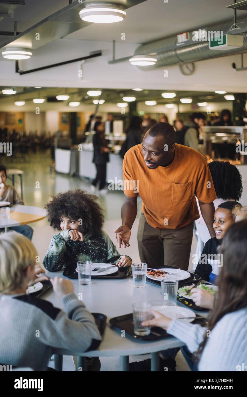 Lehrer im Gespräch mit Schülern, die während der Mittagspause in der Schulkantine am Tisch sitzen Stockfoto