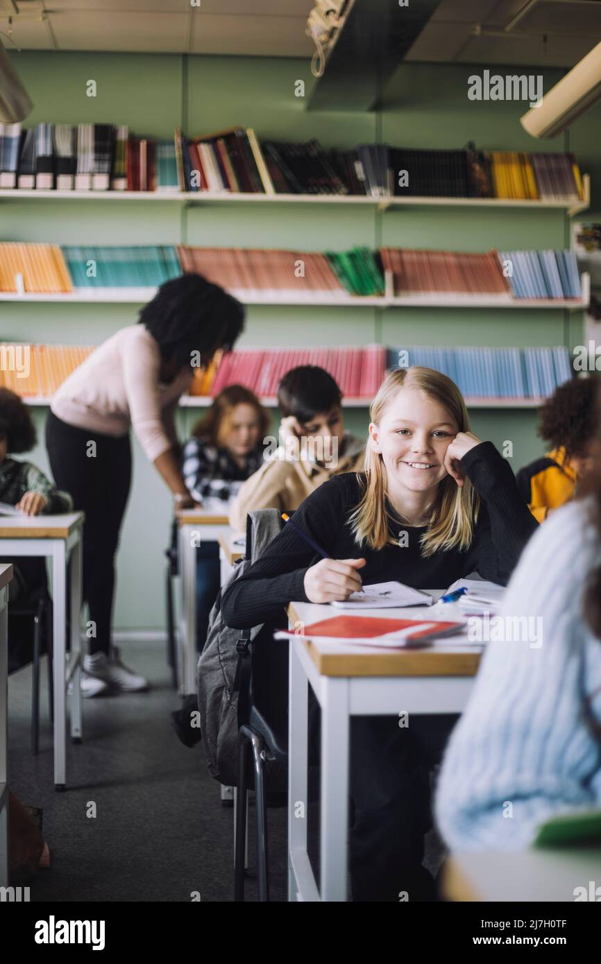 Lächelndes Mädchen, das sich an den Ellbogen lehnt, während es im Klassenzimmer am Schreibtisch sitzt Stockfoto