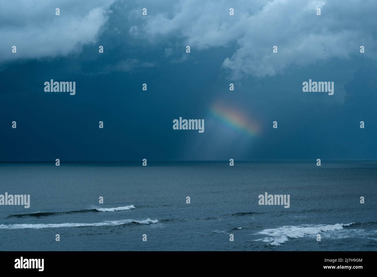 Ein kleines Stück Regenbogen vor einem schwarz-blauen Sturmhimmel mit weißen Wolken oben und dem Meer im Vordergrund Stockfoto