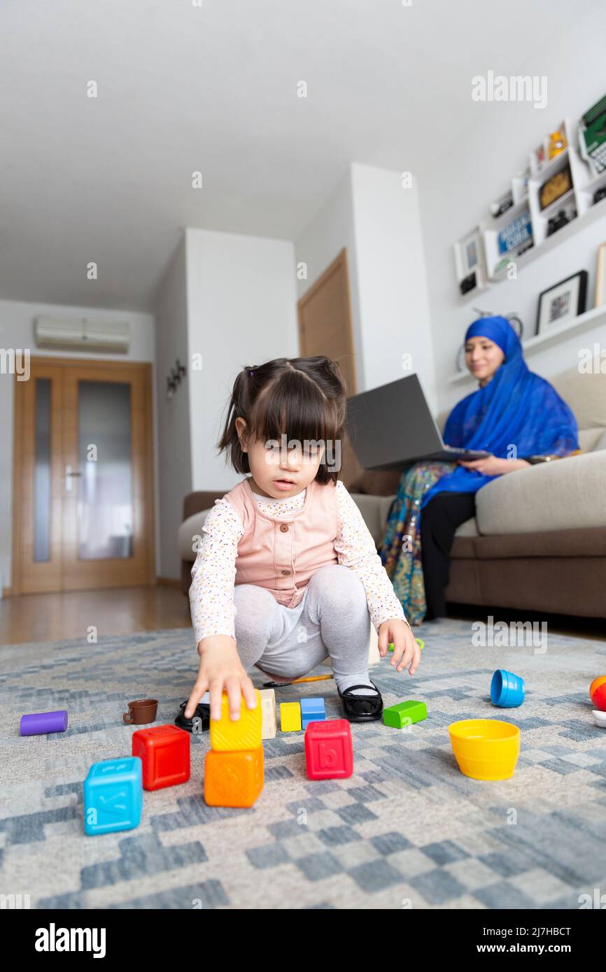 Kleines Kind spielt, während ihre muslimische Mutter von zu Hause aus mit einem Laptop arbeitet. Alleinerziehende Familie. Stockfoto
