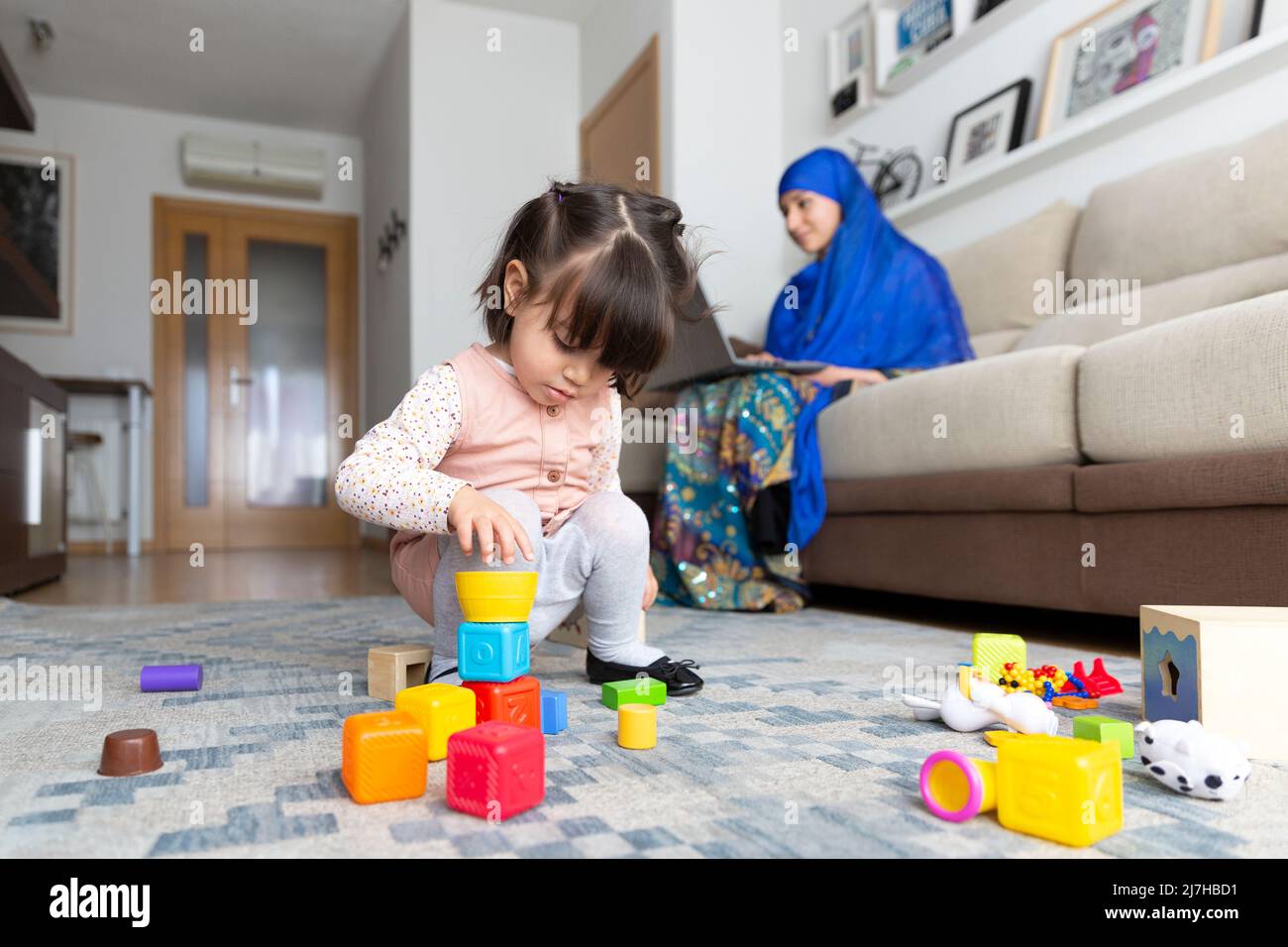 Konzept der alleinerziehenden Familie. Kleines Kind spielt, während eine junge muslimische Frau von zu Hause aus mit einem Laptop arbeitet. Stockfoto