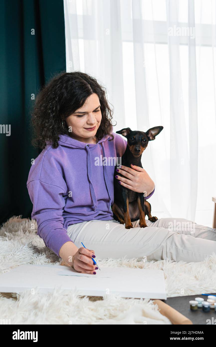 Die Erwachsene Frau, die in Innenräumen nach Zahlen malte, ihre Miniatur-Pincher in der Nähe. Personen mit Haustieren. Konzepte von Freizeitaktivitäten, Hobby und Entspannung Stockfoto