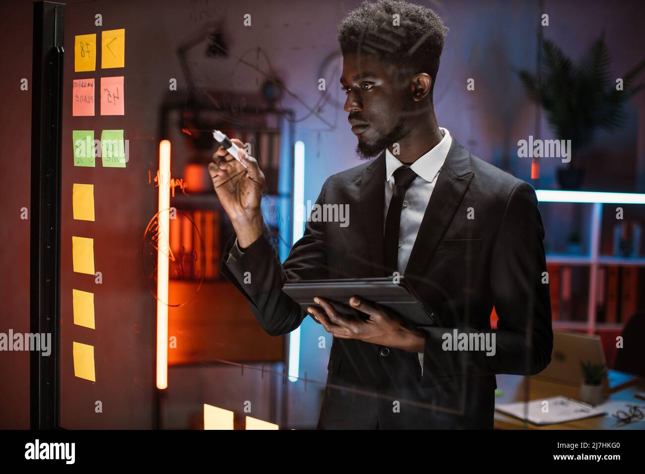 Fokussierter afrikanischer männlicher CEO schreibt mit Haftnotizen auf dem gläsernen Bürobrett und managt Projektaufgaben. Männlicher Fachmann schreibt kreative Geschäftsidee auf Glasbrett Planung Unternehmensstrategie Stockfoto