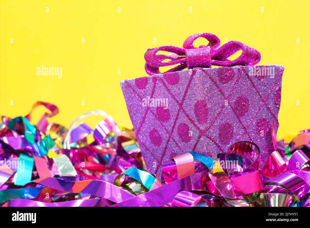 Eingewickeltes dekoratives rosa Geschenk und Schleife mit farbigen Bändern darunter auf einem gelben Hintergrund mit Kopierfläche. Stockfoto