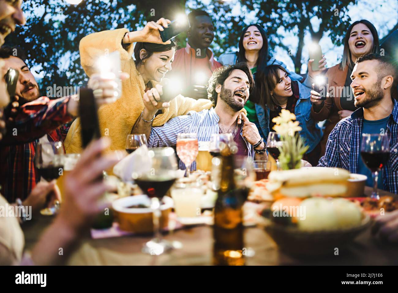 Gruppe betrunkener junger Menschen, die laut am Grilltisch singen, Wein und Bier trinken und Lichter mit Smartphones Fackeln machen - Freunde Stockfoto