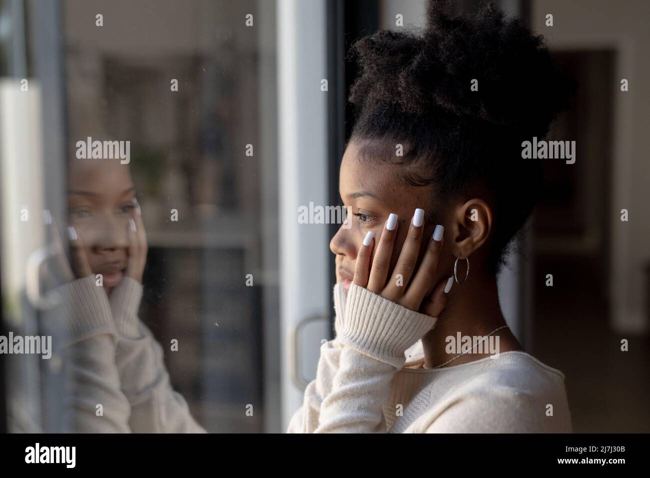 Nahaufnahme einer ernsthaften afroamerikanischen jungen Frau mit Händen am Kinn, die durch das Fenster zu Hause blickt Stockfoto