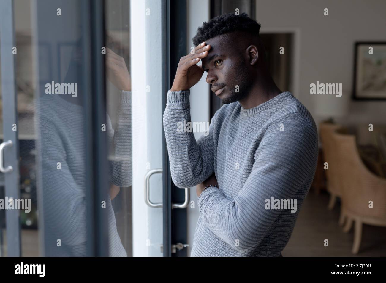 Ernsthafter afroamerikanischer junger Mann mit Kopf in der Hand und Blick durch das Fenster, während er zu Hause steht Stockfoto