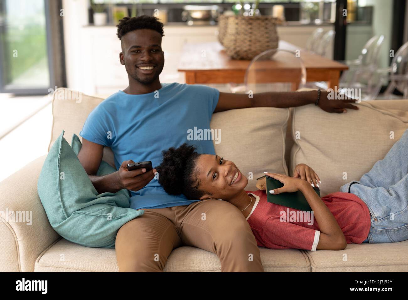Lächelnde afroamerikanische junge Frau mit Buch, das auf dem Schoß des Freundes liegt und das Smartphone auf dem Sofa benutzt Stockfoto