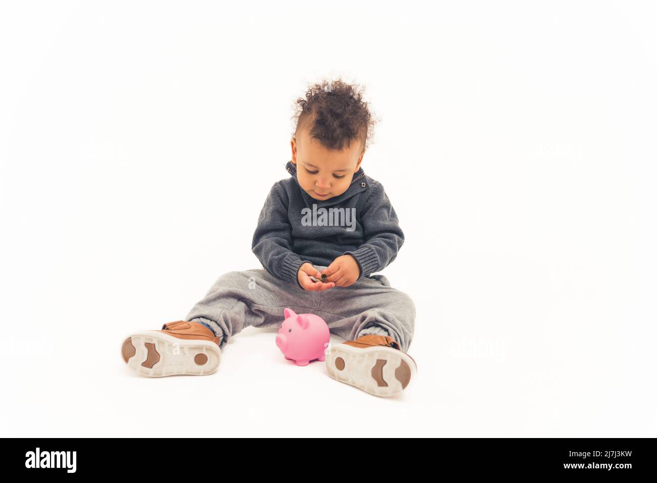 Studioaufnahme eines Biracial-Kindes, das Münzen in ein rosa Sparschwein legt, während es auf einem Boden auf weißem Hintergrund sitzt. Hochwertige Fotos Stockfoto