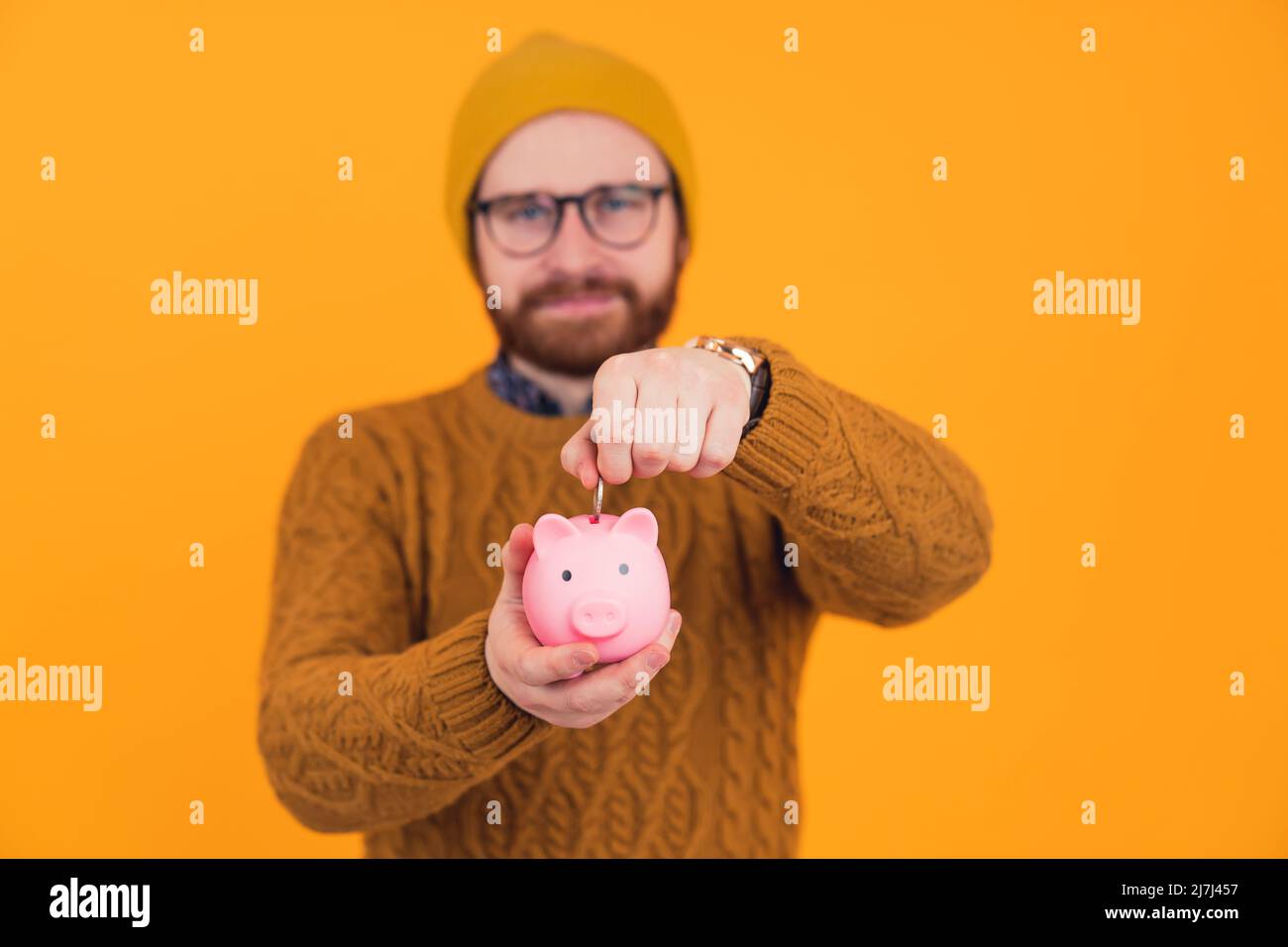 Hipster mittleren Alters europäischer hübscher Kerl in einem Studio auf orangefarbenem Hintergrund, der eine Münze in ein süßes rosa Sparschwein steckt. Hochwertige Fotos Stockfoto
