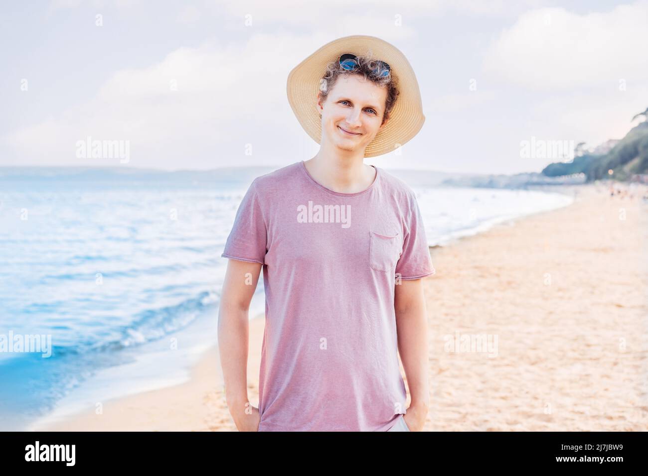 Porträt eines lächelnden jungen Mannes in einem rosa T-Shirt und Strohhut und Sonnenbrille auf seinem Kopf, der an einem sonnigen Sommertag an einem Strand am Meer steht. Entspannen Sie sich w Stockfoto