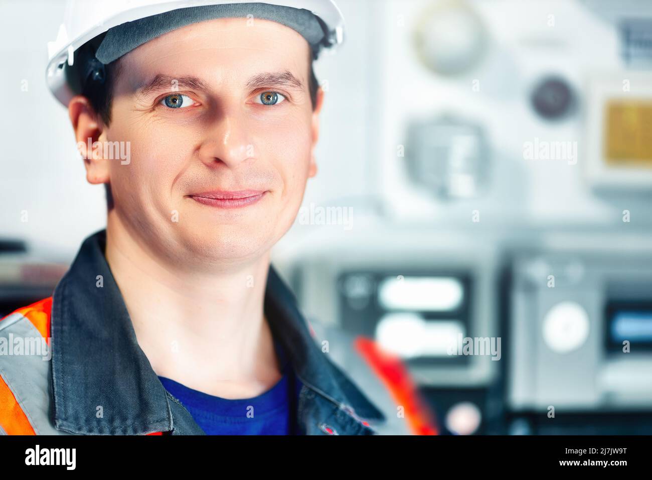 Junger positiver Ingenieur in Helm und Konstruktionsweste blickt in die Kamera und lächelt. Porträt eines jungen Schwerarbeiters im Raum mit unscharfem Hintergrund. Mitarbeiter in der Gasindustrie. Stockfoto