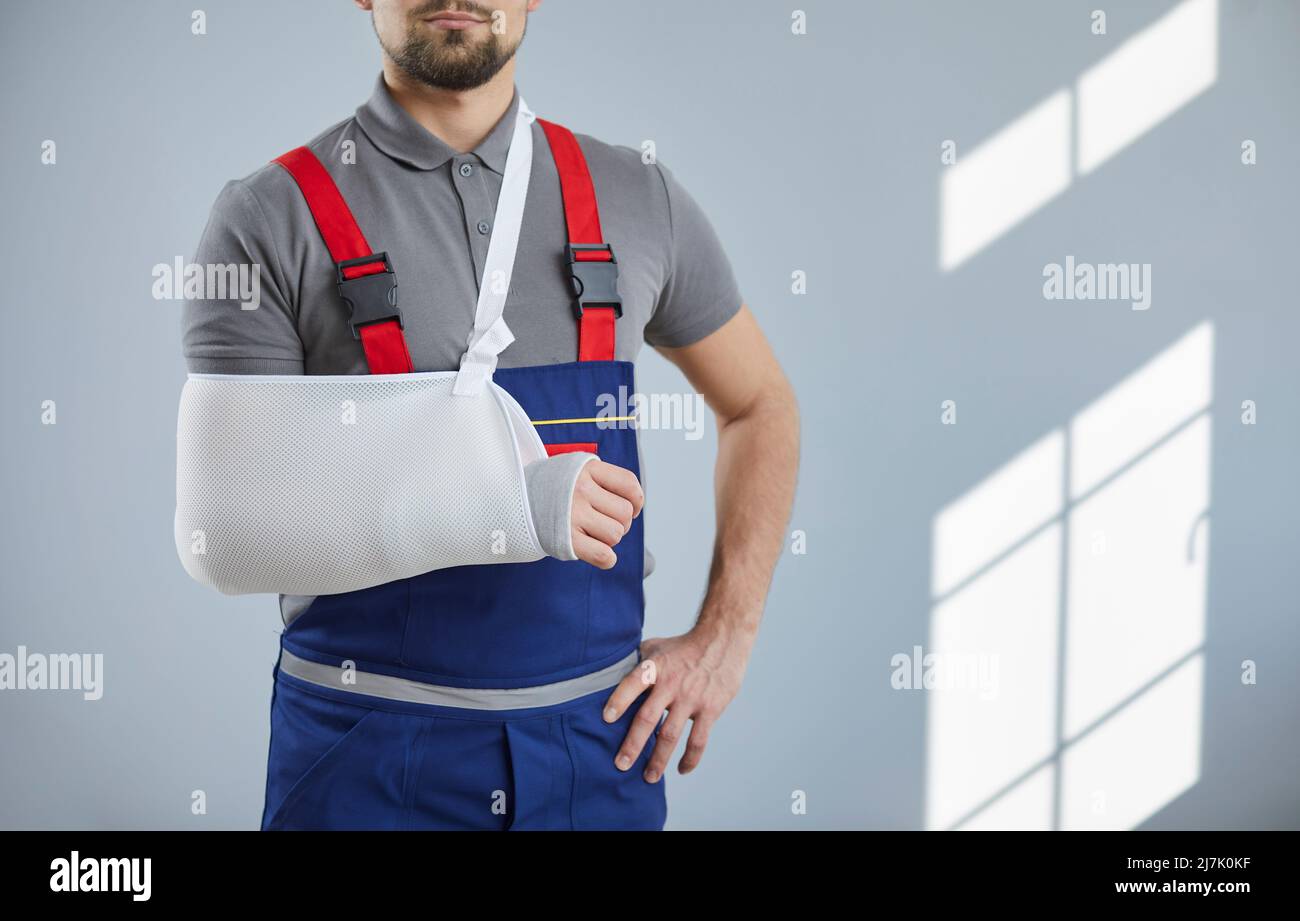 Bei der Arbeit verletzter Arbeiter steht gegen eine graue Wand mit Verband und Schlinge an seinem gebrochenen Arm. Stockfoto