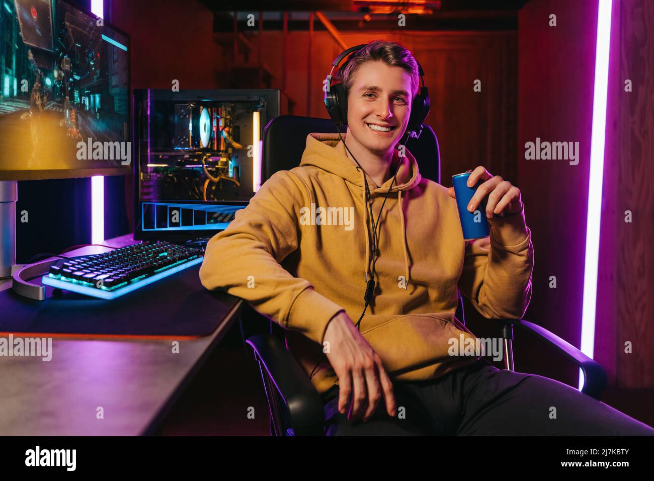 Porträt eines lächelnden Pro-Gamer, der ein Headset trägt, die Kamera anschaut, das Energie-Koffein dink hält, auf einem Spielstuhl in der Nähe eines professionellen Gaming-Setups sitzt. Cyber-Sportler spielen Shooter Computerspiel Stockfoto