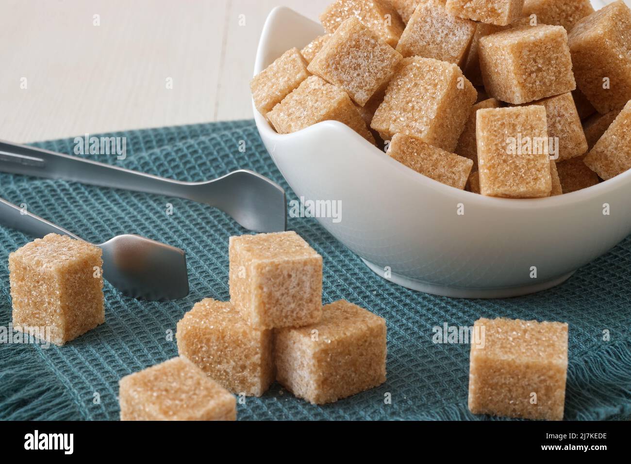 Würfelförmiger Rohrzucker in Porzellanschale auf türkisfarbener Serviette mit silberner Zuckerzange. Blick von vorne aus der Nähe, keine Leute. Stockfoto