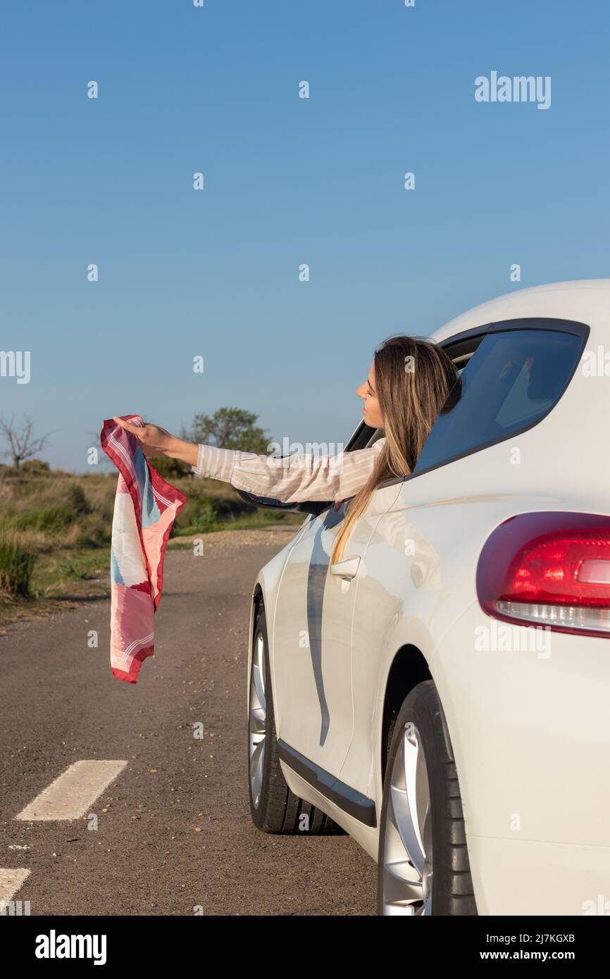 Junge Frau zieht ein buntes Taschentuch aus dem Autofenster und spürt dabei die kühle Brise auf ihrem Gesicht, während sie die Fahrt genießt Stockfoto