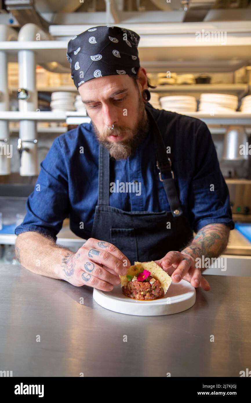 Tätowierte fokussierte männliche Koch in Schürze setzen Cracker in frischem Rindfleisch Tartare am Tisch, während der Arbeit in der Küche des Restaurants Stockfoto