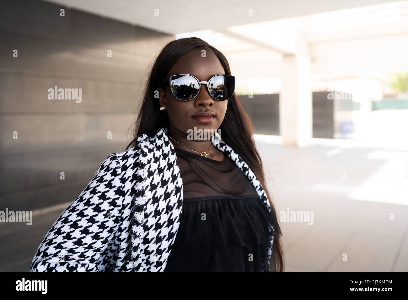 Selbstbewusste junge afroamerikanische Frau mit langen dunklen Haaren in schwarzem Outfit und trendigen Sonnenbrillen, die auf der Straße dagegen stehen Stockfoto