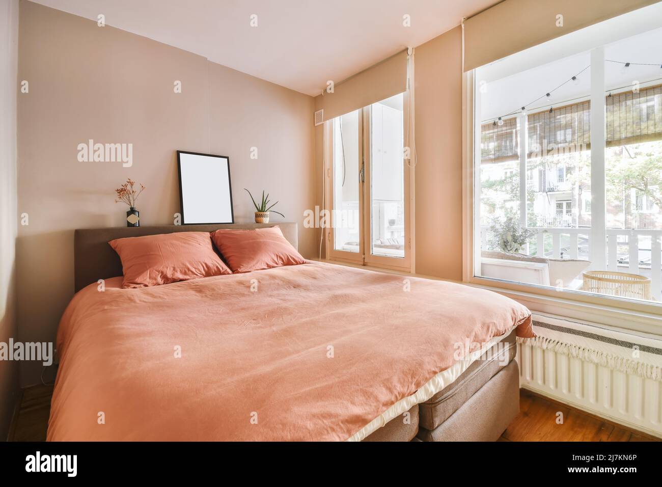 Komfortables weiches Bett mit Kissen und Decke, das in einem modernen Schlafzimmer in einem hellen Apartment neben den Fenstern platziert wurde Stockfoto