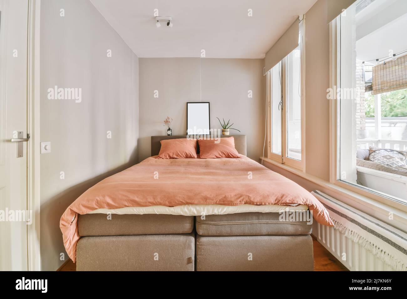 Komfortables weiches Bett mit Kissen und Decke, das in einem modernen Schlafzimmer in einem hellen Apartment neben den Fenstern platziert wurde Stockfoto