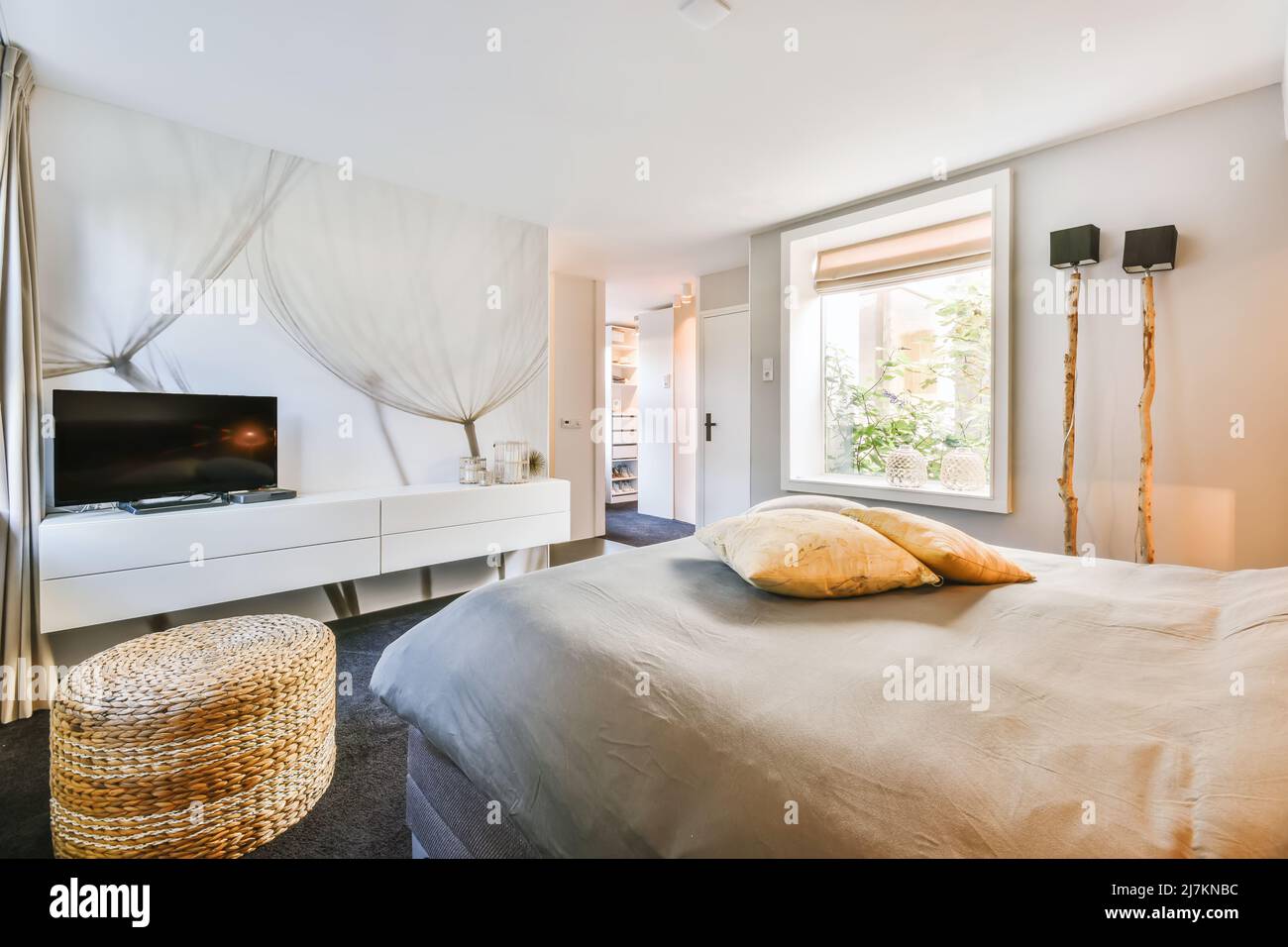Modernes Fernsehgerät auf weißem Schrank vor einem bequemen weichen Bett im geräumigen Schlafzimmer mit Rattan-Hocker und kreativen Holzlampen Stockfoto