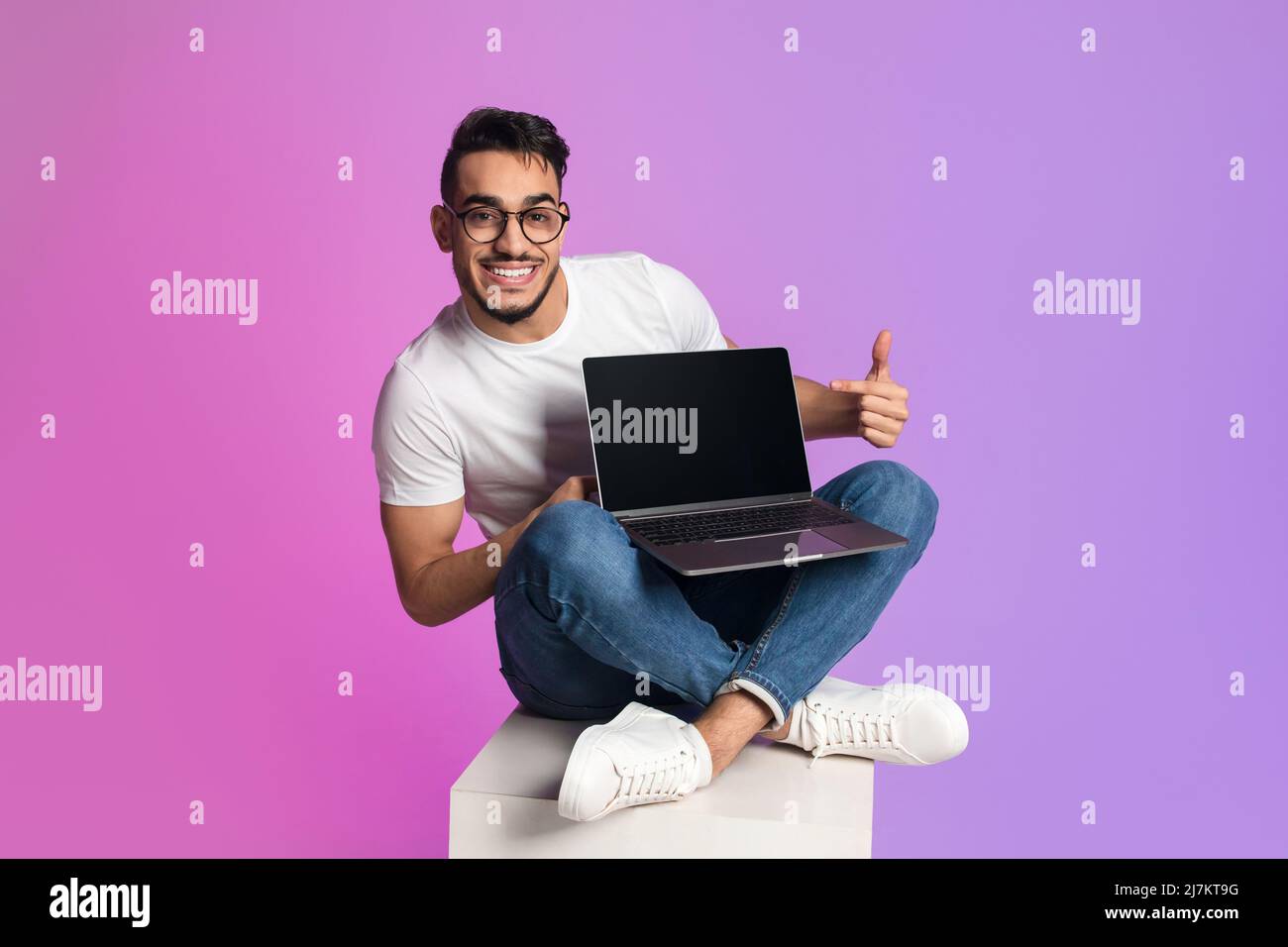 Voller glücklicher junger arabischer Mann, der mit einem Laptop-Computer überkreuzt sitzt und auf einen leeren Bildschirm zeigt und ein Modell anbietet Stockfoto