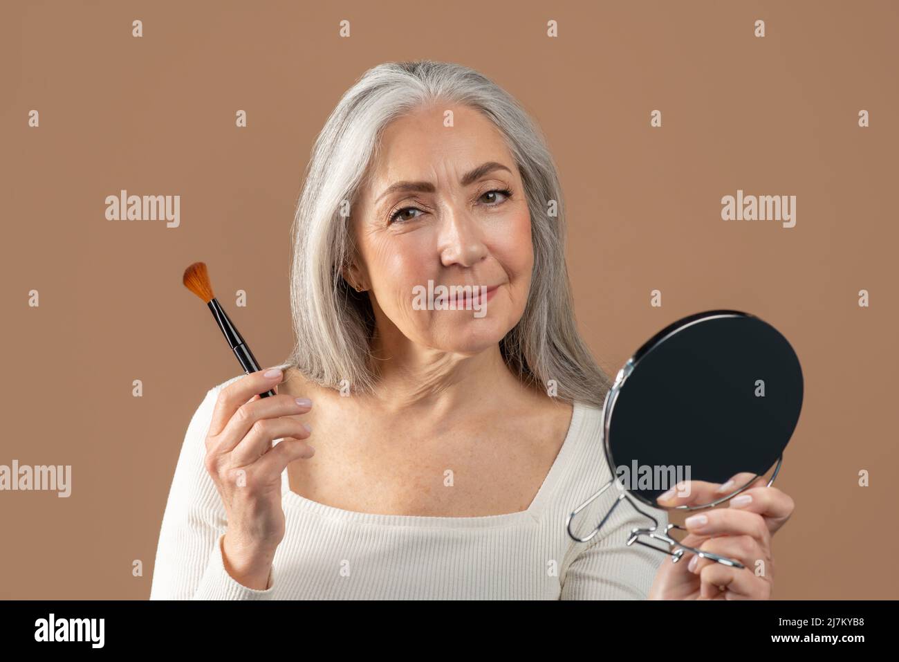Porträt einer positiven älteren Frau, die natürliches Make-up mit Pinsel aufführt und im Spiegel auf braunem Studiohintergrund schaut Stockfoto