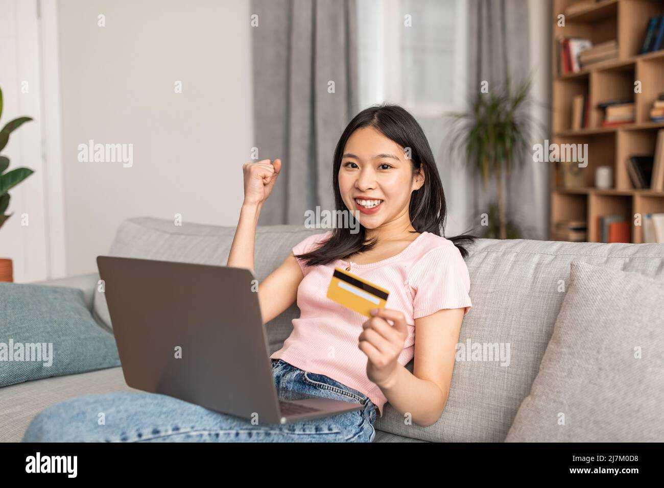 Online-Kreditkonzept. Aufgeregte koreanische Dame feiert Erfolg mit Debitkarte und Laptop, sitzt auf der Couch zu Hause Stockfoto