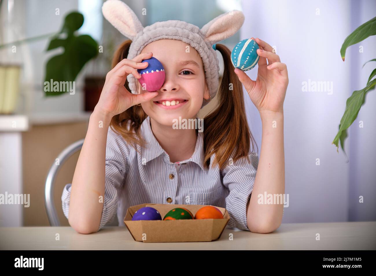 Ein positiv verspieltes Mädchen in Hasenohren hält in der heimischen Küche bunte Eier in den Händen. Frohe Ostern. Osterhintergrund. Stockfoto