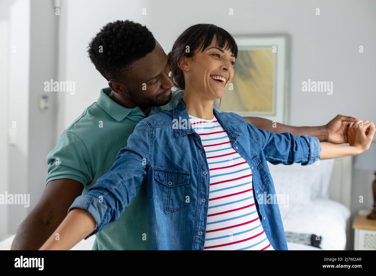 Glückliches multirassisches junges Paar mit ausgestreckten Armen, das zu Hause im Wohnzimmer steht Stockfoto