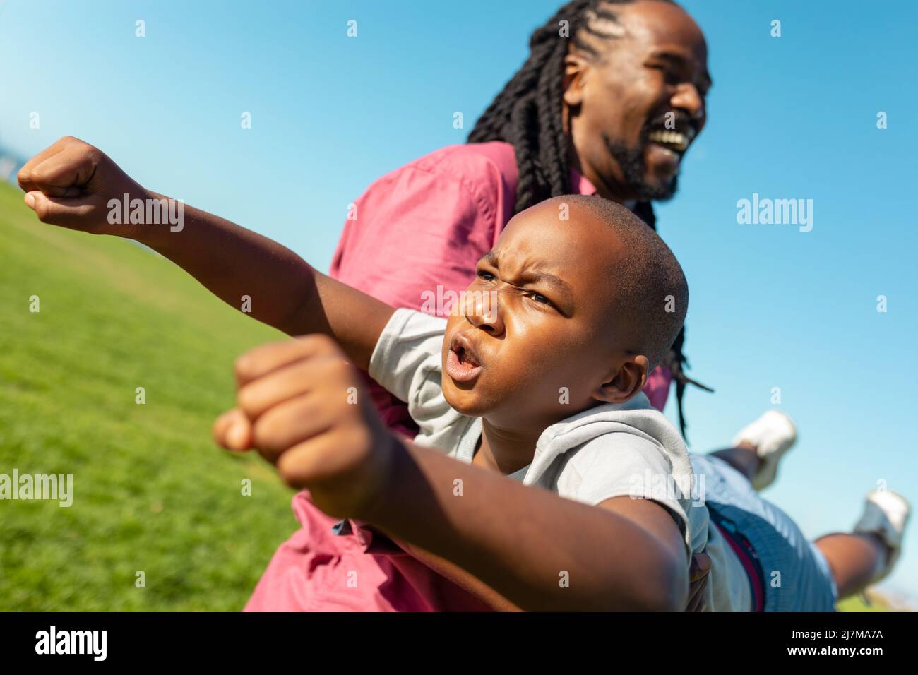 afroamerikanischer Vater trägt Sohn, während Junge imitiert als Superheld fliegen an sonnigen Tag Stockfoto