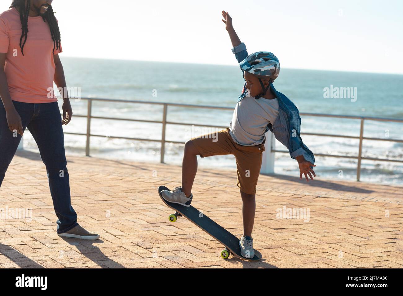 afroamerikanischer Junge, der an sonnigen Tagen dem Vater Skateboarding-Fähigkeiten auf der Promenade zeigt Stockfoto