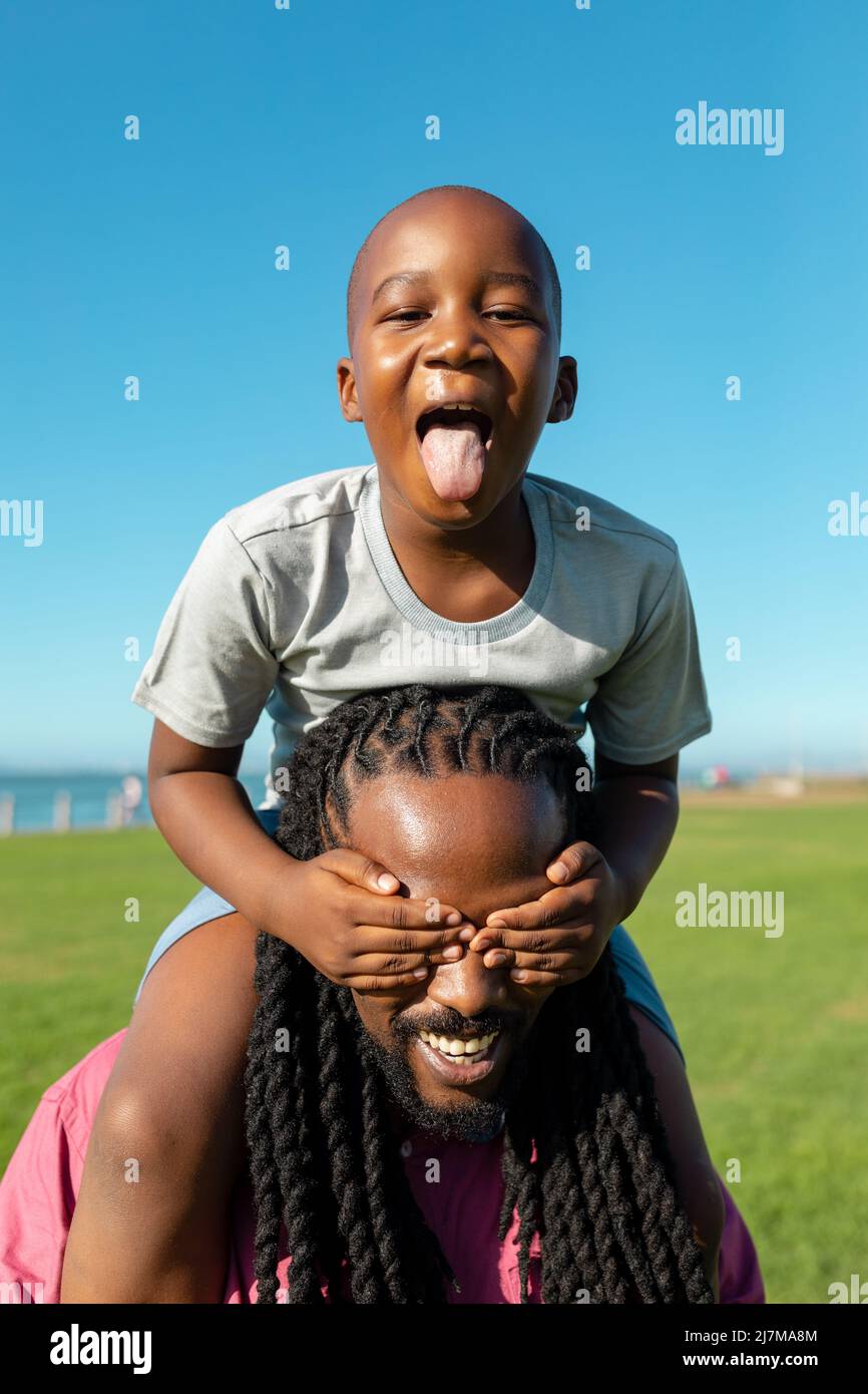 Porträt eines afroamerikanischen Jungen, der seine Zunge zeigt, während er auf den Schultern des Vaters sitzt und die Augen schließt Stockfoto