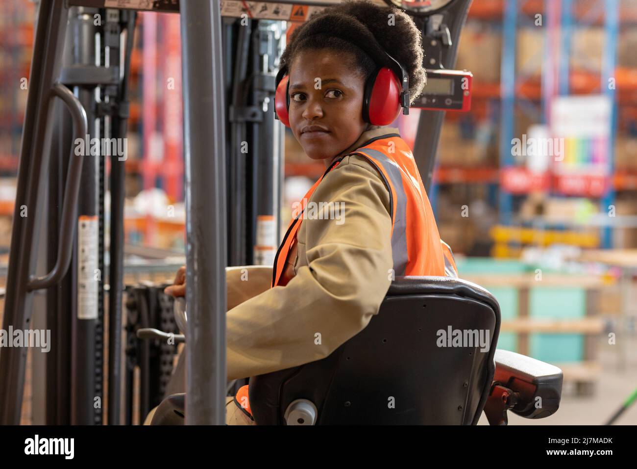 afroamerikanische junge Arbeiterin, die Ohrenschützer trägt, schaut weg, während sie im Gabelstapler sitzt Stockfoto