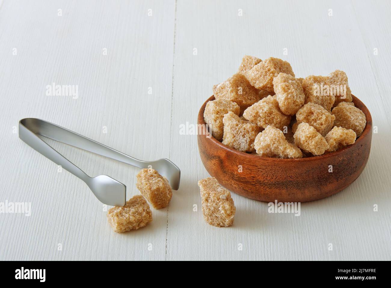 Würfelförmiger Rohrzucker in Holzschüssel auf einem weißen Holztisch mit silberner Zuckerzange. Blick von vorne aus der Nähe, keine Leute. Stockfoto